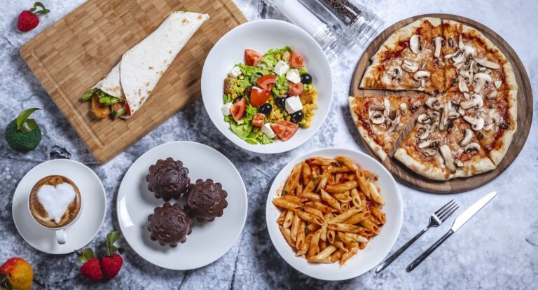 top view fast food mix greek salad mushroom pizza chicken roll chocolate muffins penne pasta and cup of coffee on the table